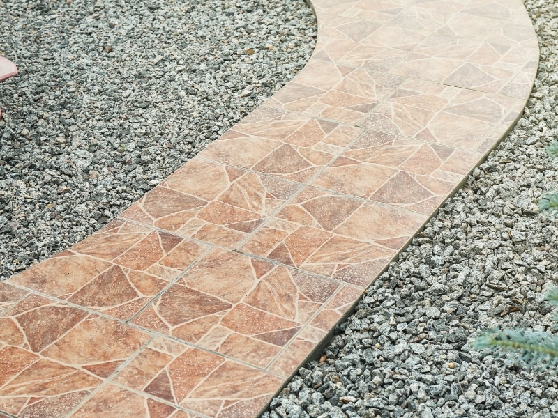 Walkway with tiles bordered by crushed stone landscaping in a garden setting.