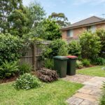 Green-lid garden organics bin beside a red-lid general waste bin with sorted garden clippings and bundled branches in a Sydney backyard.